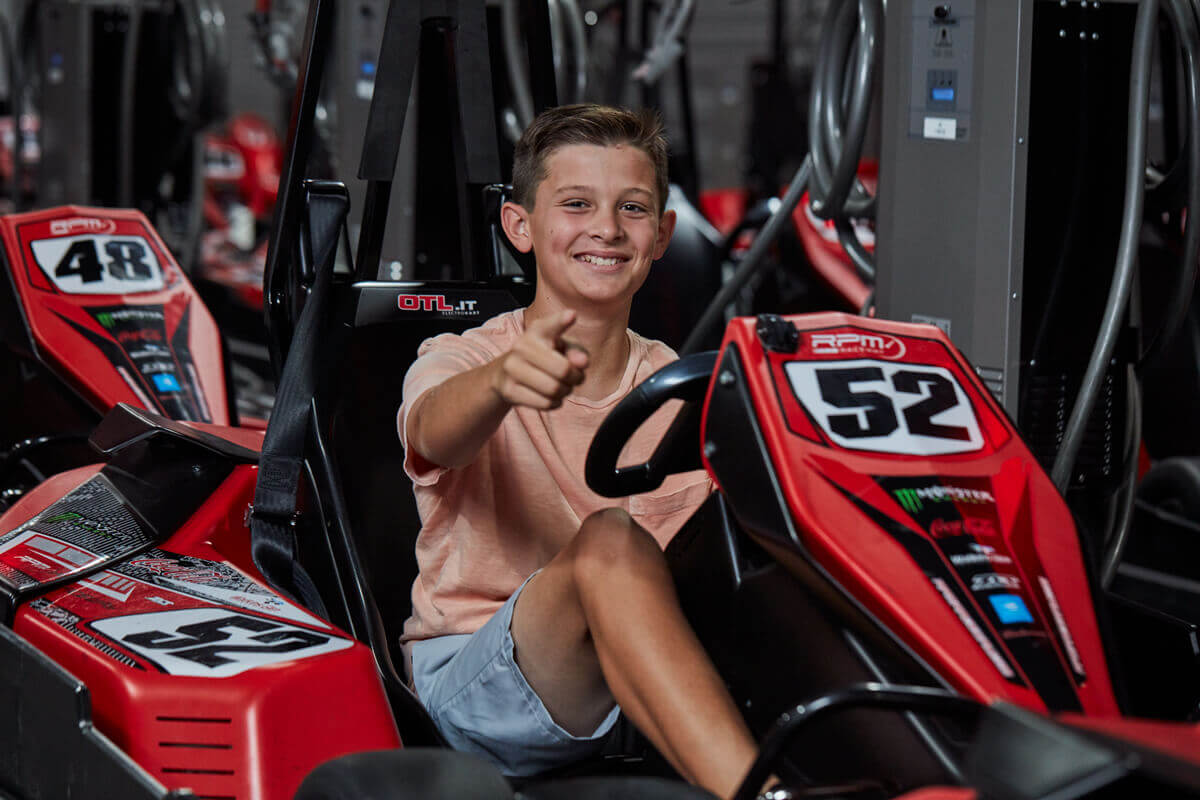 A young boy wearing a light pink t-shirt and gray shorts is sitting in a red go-kart with the number 52. He is smiling and giving a thumbs-up gesture. The background shows more go-karts with numbers, indicating an indoor go-kart facility.